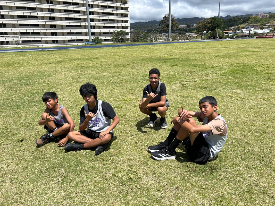Image of Sylvester, Leandro, Aeden, and Kala sitting on the grass field at the Moanalua High School track. 
